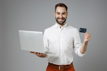 Wall Mural - Smiling young bearded business man in classic white shirt standing using laptop pc computer hold credit bank card isolated on grey color background studio portrait. Achievement career wealth concept.