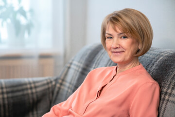 Portrait of beautiful happy positive woman, adult elderly senior mature lady sitting on couch at home in living room and looking at camera with a cheerful joyful look and smiling