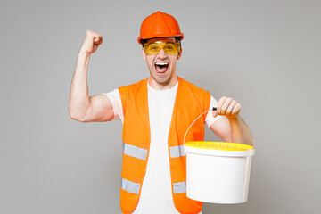 Poster - Smiling young man in orange vest protective hardhat hold paint bucket do winner gesture clench fist isolated on grey background studio. Instruments accessories for renovation apartment. Repair home