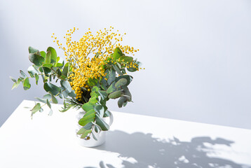 Wall Mural - a spring bouquet with yellow mimosa flowers and eucalyptus branches stands on white table with shadow. concept of 8 March, happy women's day