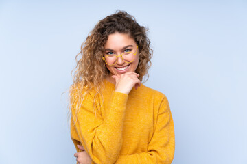 Wall Mural - Young blonde woman with curly hair isolated on blue background with glasses and smiling