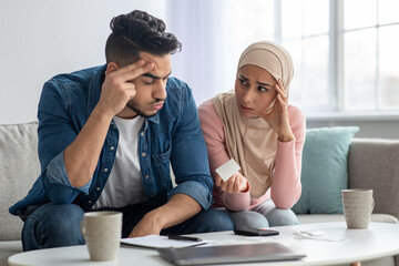 Wall Mural - Confusded lady in hijab showing bill her upset husband