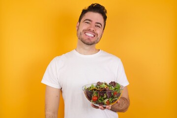 Wall Mural - young handsome Caucasian man holding a salad bowl against yellow wall laughing.