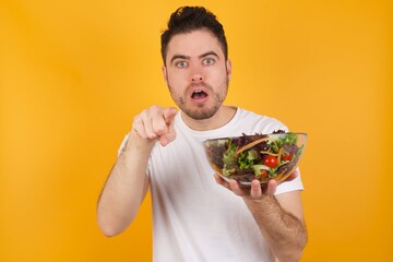 Poster - Shocked young handsome Caucasian man holding a salad bowl against yellow wall points front with index finger at camera and. Surprise and advertisement concept.