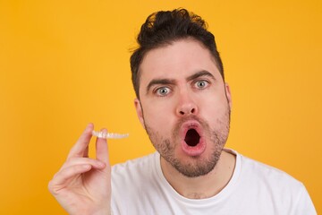 Young handsome Caucasian man standing against yellow background, holding and showing invisible aligner braces. Concept of dental health.