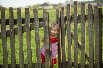 A little girl in a red dress looks out from behind a wooden fence. The child stands at the gate that leads to the garden. Cute girl in the village on a farm. Old wooden gate to the garden.