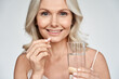 © insta_photos - Smiling happy healthy middle aged 50s woman holding glass of water taking dietary supplement vitamin pink pill isolated on white background. Old women multivitamins antioxidants for anti age beauty.