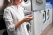 © alfa27 - Laundry, smiling woman holding phone, washing clothes via app. Cropped young caucasian female is ready to wash clothes