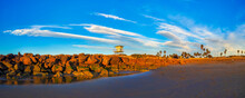 Cloud Streaked-sky On A Beach With A Breakwater And Lifeguard Tower, Birds Flying Through The Air In A Relaxing Evening Sea Shore Scene.  Golden Rocks, White Clouds And Birds Are In The Sky.
