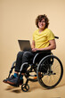 © alfa27 - handicapped person in casual wear sitting with laptop on wheelchair, isolated over beige studio background. handsome curly male enjoy working online, typing message on keyboard