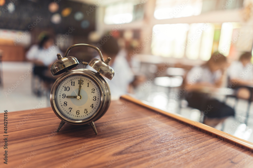 Close up vintage metal alarm clock representing lesson starting time at school, clock on teachers desk with students in the background, Asian type schooling in Thailand, timing examination testing Stock Photo |