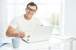 Work at ease. Latin man in white t shirt and eyeglasses smiling at camera while sitting at the table, using laptop and making notes. Businessman working from home during lockdown