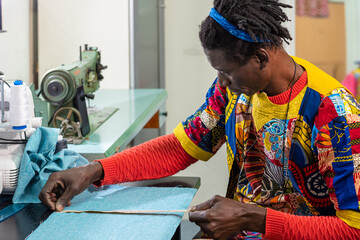 african tailor dressed in traditional clothes and fabrics at work in his workshop, production of unique clothes with modern style and colors of africa, small business, business concept of craftsman