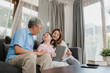 © tirachard - Asian grandparents and granddaughter using tablet at home. Senior Chinese, grandpa and grandma happy spend family time relax with young girl checking social media, lying on sofa in living room concept