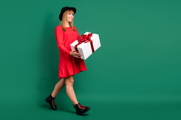 Sticker - Photo of positive cute lady hold gift box look empty space go wear hat red dress boots isolated green color background