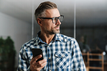 Wall Mural - Serious white-haired man in eyeglasses using mobile phone in office