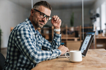 Wall Mural - Pleased unshaven man in eyeglasses working with laptop in office
