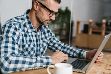 Wall Mural - Focused unshaven man in eyeglasses working with laptop in office