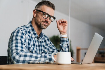 Wall Mural - Pleased unshaven man in eyeglasses working with laptop in office