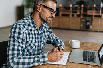 Wall Mural - Pleased grey man writing down notes while working with laptop