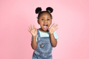 Wall Mural - Portrait of surprised screaming cute little African girl child standing isolated over pink background, looking at camera with open mouth. Concept of children's emotions, facial expression