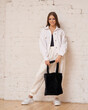 © Dmitry Tsvetkov - Young brunette teenager woman in white trousers and jeans jacket holding black textile bag and standing against white brick wall