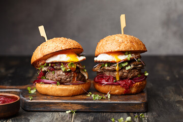 Two homemade beef burgers with mushrooms, micro greens, red onion, fried eggs and beet sauce on wooden cutting board. Side view, close up