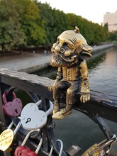 An Iron Figurine Of A Gnome Sits On The Railing Of A Bridge