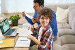 © Friends Stock - Time to study at home. Cheerful latin boy wearing protective mask while sitting at the desk together with his father, using laptop, having online lesson indoors