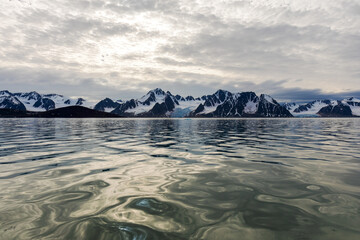 View of Raudfjorden in Svalbard with the snow-capped mountains at the horizon and the light reflecting off the ocean in the foreground.