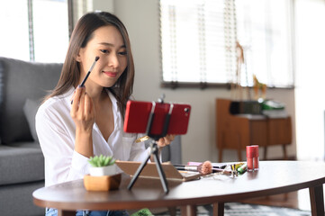 Beauty blogger influencer Asian woman using smartphone live streaming review cosmetics products in living room at home