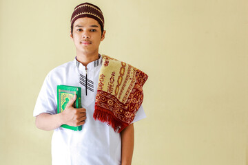 Portrait of young asian muslim holding the holy book Al-Quran and prayer mat on his shoulder get ready to pray