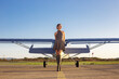 Beautiful woman in gray dress standing in front of private plane. Blue aircraft on aerodrome. Pretty model near small aircraft. Small aviation advertisement.
