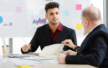 2 Adults caucasian businessmen are talking. A focused businessman is holding pen and look at a teammate and have brown cup, book, laptop put on desk and whiteboard are back with more graph on paper