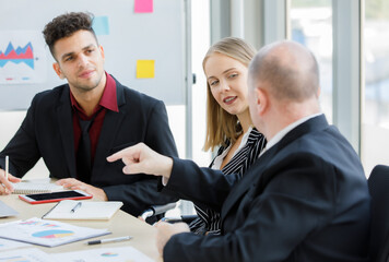 2 Adults caucasian businessmen are talking. A focused businessman is holding pen and look at a teammate and have brown cup, book, laptop put on desk and whiteboard are back with more graph on paper