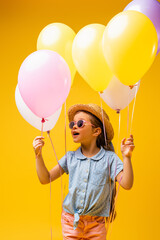 amazed kid in straw hat and sunglasses holding balloons isolated on yellow