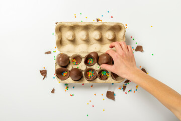 Wall Mural - Female hand takes or holds chocolate eggs and multi-colored sweet candy decorations from a tray on a light background. Easter concept, easter treats, bunny. Banner. Flat lay, top view