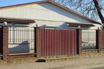 Canvas Print - red metal gates and a fence wall of black iron rods and brown bricks in front of the white wall of the house