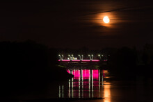 Super Full Moon In The Yellow Dark Sky With A Reflection In The River With A Colored Light Bridge, The Moonlight Reflects The Surface Of The Water. Long Exposure