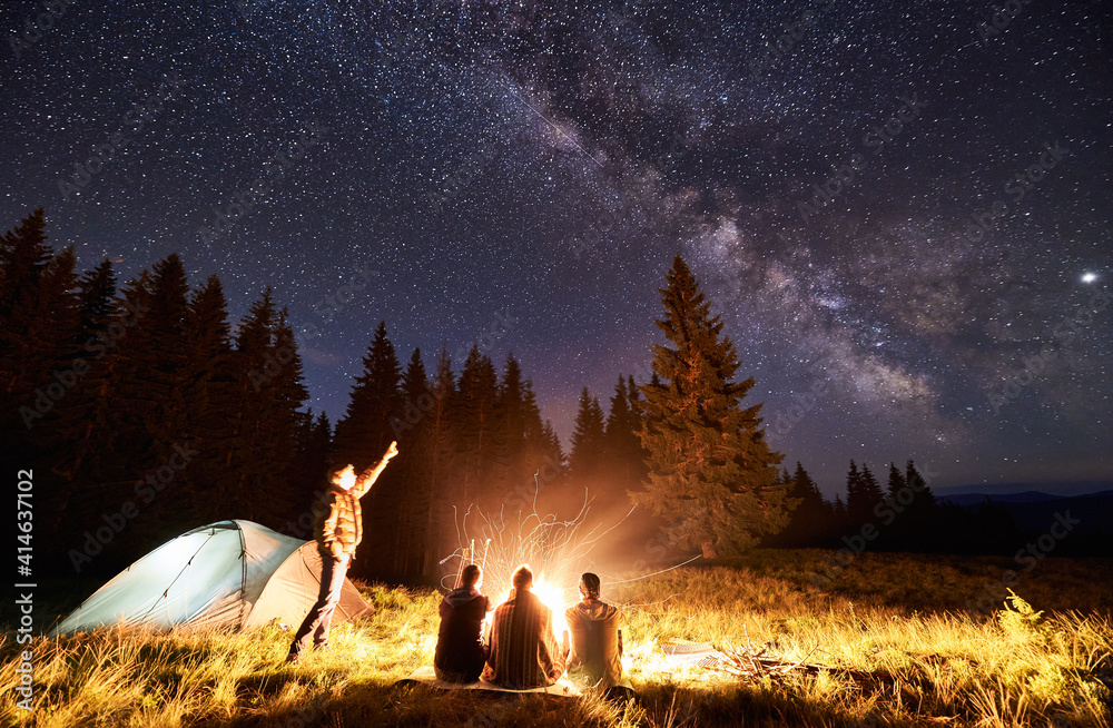 Male showing his friends Milky Way over camping. Guys are sitting by the campfire on the background of the valley of mountains and pine forest, enjoying the evening starry sky. Outdoor recreation - obrazy, fototapety, plakaty 