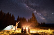 © anatoliy_gleb - Male showing his friends Milky Way over camping. Guys are sitting by the campfire on the background of the valley of mountains and pine forest, enjoying the evening starry sky. Outdoor recreation