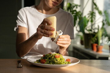 Close up of woman hands grating Parmesan cheese in pasta with sauce pesto, fresh cherry tomatoes, living room on background. Homemade Italian cuisine.