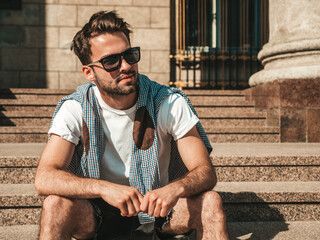 Portrait of handsome confident stylish hipster lambersexual model.Man dressed in white T-shirt. Fashion male sitting at the stairs on the street background in sunglasses