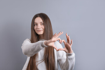 Wall Mural - Portrait of a smiling young attractive woman showing heart gesture with two hands and looking at camera isolated over grey background