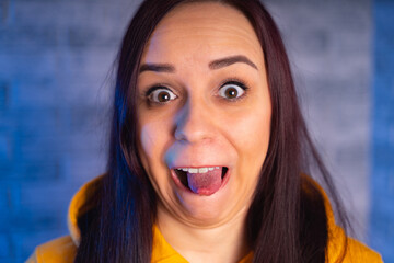 Wall Mural - Portrait of young woman with grimacing face. Close up of crazy female in yellow hoodie monkeying on gray brick background.