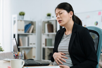 Wall Mural - asian japanese pregnant woman in suit sitting in office chair and touching big belly. illness maternity lady worker with stomach ache. sick future motherhood hand holding painful abdomen in workplace