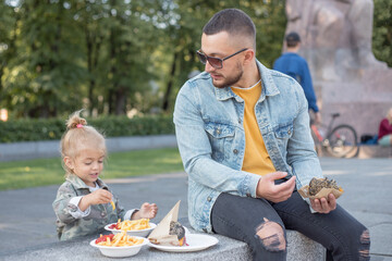 Father and daughter are eating burger and french fries on the street in the park, Man and little girl enjoying fast food.