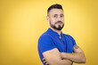 Handsome man with beard wearing blue polo shirt over yellow background looking to the side with arms crossed convinced and confident