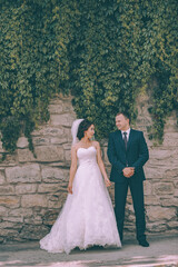 Wall Mural - Wedding couple poses directly in front of the camera against the background of a stone wall with ivy. The bride is dressed in a classic wedding dress and the groom in a dark blue suit.