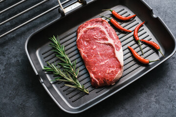 Wall Mural - Raw rib eye beef steak on black grilling pan with chili pepper and rosemary, top view.